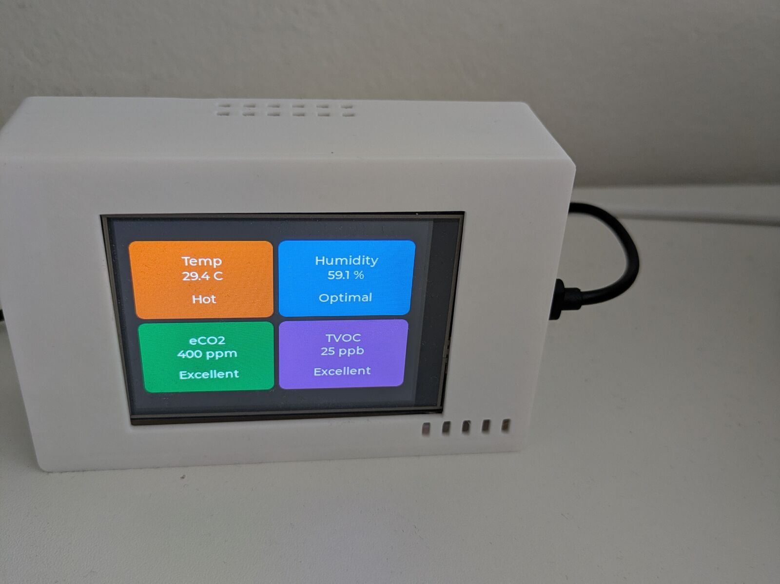 A white 3D-printed box standing on a desk with a USB cable plugged into its side. The screen shows four colored cards: Temp 29.4 C Hot, Humidity 59.1 % Optimal, eCO2 400 ppm Excellent, TVOC 25 ppb Excellent. Vent slots are visible on the top wall and next to the screen.
